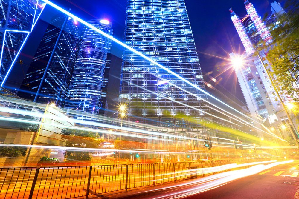 Avenida movimentada à noite com rastros de luz entre edifícios corporativos